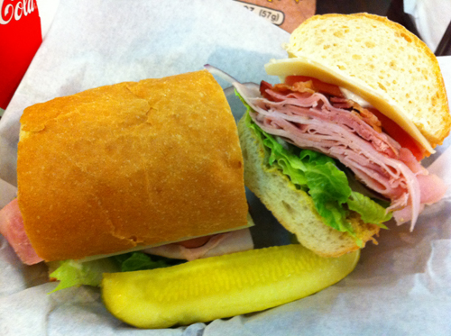 A picture-perfect deli-sliced ham sandwich. Three types of ham piled high, bacon, provolone and accoutrement on a white roll. A pickle spear supports the composition.